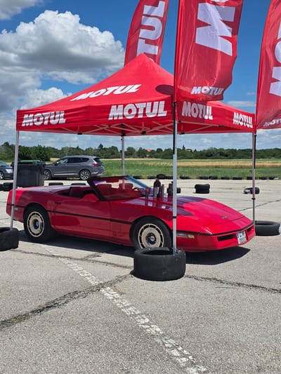 A red convertible Chevrolet Corvette is parked under a red pop-up canopy with "MOTUL" branding on it, at an outdoor event. The car is positioned on an asphalt surface, likely a racetrack or parking lot, with tire marks visible on the ground. Several large black tires are placed around the car, possibly as markers or barriers. In the background, a grey SUV and black trash bins are visible, along with a line of trees and a cloudy blue sky. The scene appears to be a promotional event or a pit stop area for a car gathering. The location is identified as Akatsievo, Bulgaria.