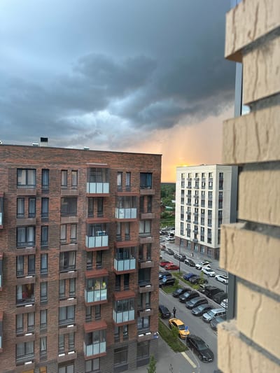 The image shows a street view in Laikovo, Russia, with two multi-story residential buildings prominently featured. The building on the left is constructed with dark red brick, and features numerous windows and balconies. The building on the right is lighter in color, possibly white or light beige, and also has many windows and balconies. Between and around the buildings, a street is visible with several parked cars of various makes and models, including black, silver, white, and yellow vehicles. A single individual, a person, is seen walking on the sidewalk next to the parked cars. The sky above is dramatic, with heavy, dark clouds suggesting impending rain or a recent storm, with a sliver of sunset visible on the horizon behind the lighter building, casting a warm glow. The foreground is partially obscured by a beige brick wall, likely part of another building or balcony. The overall scene depicts an urban residential environment under a moody sky.