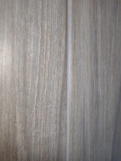 The image shows a close-up view of a textured surface, likely wood veneer or laminate, with a vertical seam running down the center. The texture displays fine, parallel lines and variations in shade, creating a wood grain effect. The colors are predominantly muted grays and light browns. The lighting casts subtle shadows, highlighting the texture and the slight indentation of the seam. The scene appears to be an interior surface, possibly a wall panel, cabinet door, or furniture. There are no people, animals, or any identifiable landmarks that would place this image in Trudobelikovskiy, Russia.