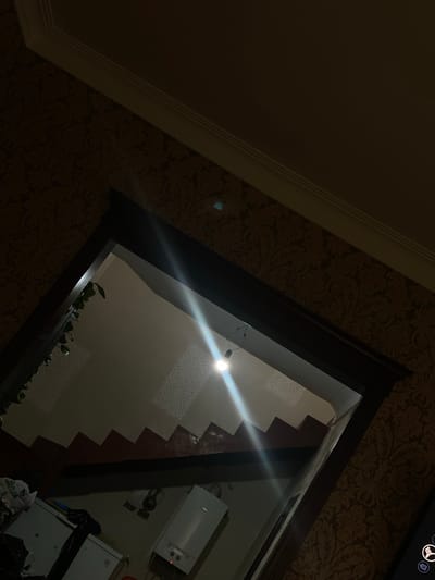 The image shows an interior scene, likely a room with patterned wallpaper. A large mirror or reflective surface dominates the right side of the frame, angled to show a ceiling with a bright light source creating a glare. Inside the reflection, we can see part of a white appliance, possibly a water heater or a small refrigerator, with some red indicator lights. There is also a dark object resembling a lamp and some bags of items. The overall lighting is dim, suggesting it might be evening or a room with limited natural light. A plant with green leaves is visible on a shelf or ledge within the reflected area. The left side of the image shows a ceiling corner with molding. The scene appears to be in Bogatyryovka, Russia. There is no clear indication of people or specific activity, but the presence of appliances and storage items suggests a domestic or utilitarian space.