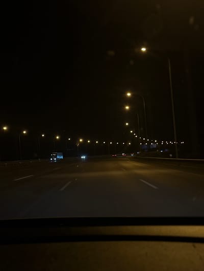 The image shows a nighttime view from inside a vehicle on a highway. Streetlights illuminate the road, which has white lane markings. Several vehicles are visible, including a truck and cars. The environment is dark, with the lights from the street and vehicles being the primary sources of illumination. The location is identified as the A-2 highway in San Fernando de Henares, Spain. The view is from the perspective of a driver or passenger, looking forward along the highway.