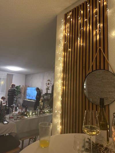 The image shows a party scene reflected in a round mirror hanging on a wall adorned with horizontal wood slats and string lights. A partially visible table holds wine glasses, a bottle of white wine, and other items. The reflection reveals people gathered around a television and dining table in a brightly lit room decorated with festive elements.
