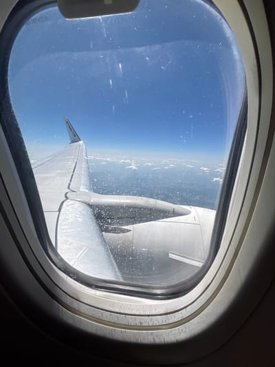 This media file is a photograph taken from an airplane window. The subjects are the airplane's wing and engine, visible through the oval-shaped window. The scene is the sky and clouds from high altitude, with glimpses of land below.

The image captures the view during a flight. The time of day appears to be daytime, with bright sunlight illuminating the scene. The weather looks clear with some scattered clouds, indicating good flying conditions. The presence of the Ryanair logo on the wing's tip suggests the flight is operated by Ryanair. Although the exact location is not explicitly visible from the aerial view, the prompt provides context that the photo is related to Majdalena, Czechia, implying this might be the region over which the plane is flying or a destination. There are no people visible in the image, and no specific emotions can be inferred.

Notable details include the texture of the airplane's wing and engine, with some minor dirt or water droplets on the surface of the window and wing. The distinct shape of the airplane window frame is also a prominent feature. The text "RYANAIR" is clearly visible on a small flag attached to the wingtip. The orientation of the photograph is unusual, appearing to be taken at an angle or possibly indicating the plane is banking or flying sideways relative to the viewer's perspective.