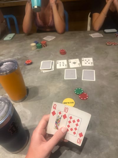 A poker game is in progress on a gray surface. The community cards include a 9 of diamonds, Ace of spades, 6 of diamonds. Player's hand reveals a 10 and Queen of diamonds. Various colored poker chips are scattered around the table, along with a "Big Blind" marker. Two people are seated around the table, and a drinking cup is in the foreground. The lighting appears dim.