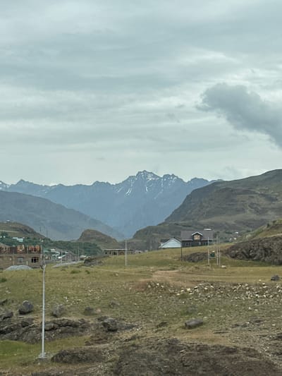 The image depicts a cloudy, overcast sky over a mountain landscape in the city of Dras, India. In the foreground, a rocky, grassy terrain is visible, with a few scattered boulders and a tall, slender pole. Further back, there are several buildings under construction, appearing as concrete structures with exposed framing and empty window openings. Beyond these, a small settlement with houses can be seen nestled on a hillside. The middle ground is dominated by rolling hills covered in sparse vegetation. In the distance, a majestic mountain range with snow-capped peaks stretches across the horizon. The overall impression is one of a remote, developing area within a rugged, mountainous environment.