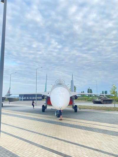 In Arablyar, Russia, an outdoor display features a parked jet fighter aircraft prominently in the foreground. The jet's nose is pointed towards the viewer, with its cockpit canopy visible. To the left of the jet, another aircraft's tail is partially visible. In the background, a tank is parked on a grassy area, and a low building with dark siding and numerous windows is situated behind the jet. Several lampposts are also present in the scene. Two individuals are seen near the jet fighter. One person, wearing dark clothing, is standing near the jet's nose gear. Another person, also in dark clothing, is standing further back near the jet's wing. The ground is paved with interlocking bricks arranged in a pattern of light and dark gray. The sky above is cloudy.