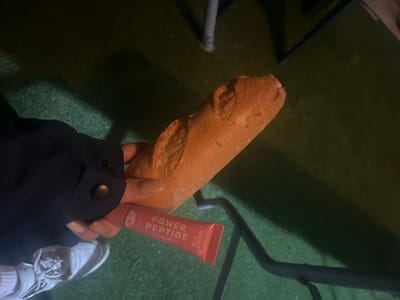 A dimly lit, close-up shot captures a person's hand holding a baguette and a cosmetic tube in Pobeda, Bulgaria. The scene appears to be an outdoor or semi-outdoor setting, characterized by a dark green, textured ground surface, possibly artificial turf or a dimly lit carpet, and a dark metal furniture leg visible in the background. The low light suggests it is nighttime or late evening.
A person's hand, adorned with long, light-colored manicured nails, prominently holds a light-brown, crusty baguette. Just above the hand, a peachy-pink tube of cosmetic product is also held. Visible text on the tube identifies it as a "trend it up" product, specifically "POWER PEPTIDE" with a "COTTON CANDY" designation, and also indicates "SPF 30." The person's arm is covered by a dark blue or black jacket sleeve featuring a black snap button. In the upper right corner of the frame, a section of a white sneaker is visible. The image focuses on these items, presenting a casual snapshot with a dark and slightly indistinct background.