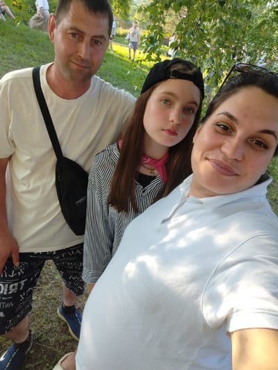 Three people are captured in a selfie outdoors, likely at Detskii Dom (Bashenino), Russia. A man in a white t-shirt and dark patterned shorts is on the left, with a black crossbody bag. Beside him is a young woman wearing a black baseball cap, a black and white striped long-sleeved shirt, and a pink scarf. In the foreground, on the right, is another woman wearing a white polo shirt, smiling at the camera. She has sunglasses perched on her head. The background shows a grassy area with trees and dappled sunlight, suggesting a park or recreational area.
