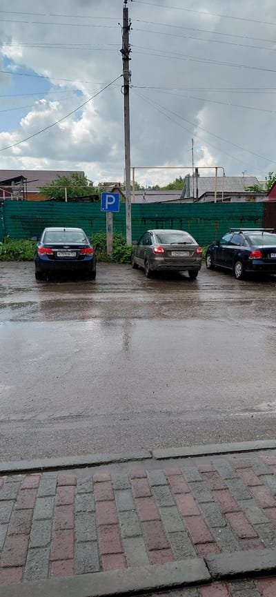 The image displays an outdoor street or parking area scene in Agapovka, Russia, under an overcast sky with visible patches of blue. The ground consists of wet asphalt, indicating recent rainfall, which reflects light and surrounding objects.
In the immediate foreground, a sidewalk constructed from alternating grey and reddish-brown rectangular paving bricks is present, separated from the road by a dark grey concrete curb.
The midground is occupied by three sedans parked on the wet asphalt, all facing away from the viewer. From left to right: a dark blue Chevrolet Cruze bearing Russian license plate "M759OB 177" (Moscow region code); a brown/grey Lada Granta with Russian license plate "T240BO 77" (Moscow region code); and a partially visible dark blue Volkswagen Polo, equipped with a roof rack, with a partially obscured Russian license plate showing "P2". Adjacent to the Chevrolet and Lada, a blue rectangular parking sign with a white "P" is mounted on a thin grey pole. Green vegetation, including weeds and bushes, grows at the base of this pole and along the adjacent fence.
Behind the vehicles, a green corrugated metal fence, displaying wear and some rust, spans horizontally across the scene.
The background features several buildings, likely residential or utility structures, with brown or reddish roofs (some tiled, some corrugated). Various wooden frameworks and sparse green trees are also visible behind the fence and structures. A prominent tall concrete utility pole, equipped with multiple cross arms, insulators, numerous electrical wires, and attached utility boxes, rises from the midground into the sky on the right side of the image. A yellow/beige metallic railing or frame is visible in front of some background buildings, to the right of the main utility pole.
The sky is predominantly grey and cloudy, though a patch of clearer blue sky with white cumulus clouds is visible in the upper left quadrant. No individuals are present in the depicted scene.