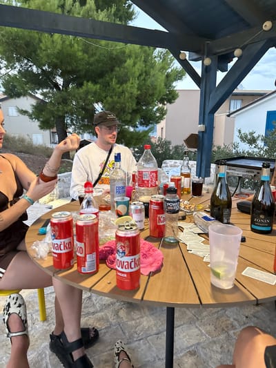 This image depicts an outdoor social gathering under a wooden pergola in Betina, Croatia.
**People:**
Three individuals are partially visible around a circular wooden table.
1. On the left, a fair-skinned female, largely cropped, wears a brown top. Her left arm is raised with the hand open, adorned with multiple bangles. Her feet, wearing black-and-white patterned slip-on shoes, are visible beneath the table.
2. In the center, a fair-skinned male wearing a white long-sleeved shirt with a colorful graphic, dark-rimmed glasses, and a dark baseball cap, looks towards the left. A black bag strap crosses his torso.
3. In the bottom right, legs and feet, one pair in dark sandals and another in black-and-white patterned slip-on shoes, are partially visible, indicating at least one more person present.
**Subjects/Objects:**
The wooden table is laden with numerous items, primarily beverages:
* Three red aluminum cans of "Kovačičko LAGER," one with a lemon slice on top.
* One large plastic bottle of Coca-Cola.
* One clear glass bottle labeled "COSMOPOLITAN POLITAN Vodka."
* Two dark glass bottles of prosecco/wine, one clearly labeled "ZONIN PROSECCO."
* A clear plastic bottle labeled "POWER ROD."
* A small red plastic bottle and a brown glass bottle.
* Various drinking vessels including a tall clear plastic cup with a lime wedge, a multi-colored tumbler, and other glasses.
* Other items on the table include a stack of white playing cards, a pink fabric cloth, loose plastic wrappers, and what appears to be cigarette butts in a small white dish.
**Environment:**
The setting is an outdoor patio or terrace with a light-colored paved floor. A dark wooden pergola structure, equipped with string lights (white globes), provides overhead cover. In the background, lush green pine-like trees are prominent, behind which several residential buildings with light-colored facades are visible. One building features a distinctive blue shuttered window. To the far right, a flat-top grill sits on a stone surface. The sky appears overcast.