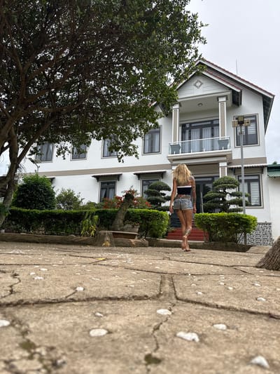 A woman with blonde hair and tattoos is standing barefoot on a paved walkway in front of a white, multi-story building with gray trim and dark windows. She is wearing a black tank top and denim shorts. The building appears to be a house or hotel. There are trees and manicured bushes in front of the building, and a large tree branches out from the left side of the frame. The walkway is made of concrete pavers and has some small white pebbles scattered on it. The scene is outdoors, and the sky is overcast, suggesting it might be a cloudy day. The woman's posture is relaxed, and she appears to be looking away from the camera. The overall setting is residential or hospitality-focused, possibly in Thôn 2, Vietnam, given the location context. No text is visible in the image.