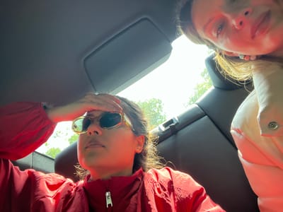 Two people are inside a car. The person in the foreground wears a red jacket and sunglasses, holding their hand to their forehead. The other person is partially visible in the upper right corner. The car interior is dark gray, with a sun visor visible in the upper left. The location cannot be determined from the image.