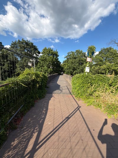 The image shows an outdoor scene on a sunny day, with a clear blue sky and some fluffy white clouds. The perspective is from someone looking down a paved walkway that curves to the right. The walkway is bordered on the left by dense green foliage and a wire fence. On the right, the walkway is bordered by more greenery, with a shadow of a person and a fence cast across the paving stones.
In the background, there are trees and a signpost with some text on it, partially obscured by vines. The sign appears to be blue and white, with some red elements. The shadows suggest it is likely late morning or early afternoon.
The overall impression is of a pleasant, sunny day in a semi-urban or park-like setting. The presence of the fence might indicate some ongoing construction or a boundary. The shadow of the person suggests they are taking a selfie or holding up a phone to capture the scene. There are no specific people clearly visible, only a shadow. The location is identified as Dorsten, Germany.