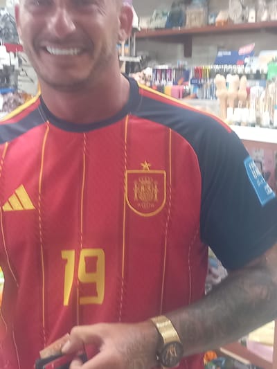 The image shows a man wearing a red and yellow striped Spain national football team jersey. The jersey has the Adidas logo on the left chest and the Spanish crest on the right chest. The number "19" is visible on the front of the jersey. The man has a gold watch on his left wrist and tattoos on his right arm. He is smiling. The background appears to be a retail store, with shelves stocked with various items. The exact location within the city of Kos, Greece, cannot be determined from the image alone.