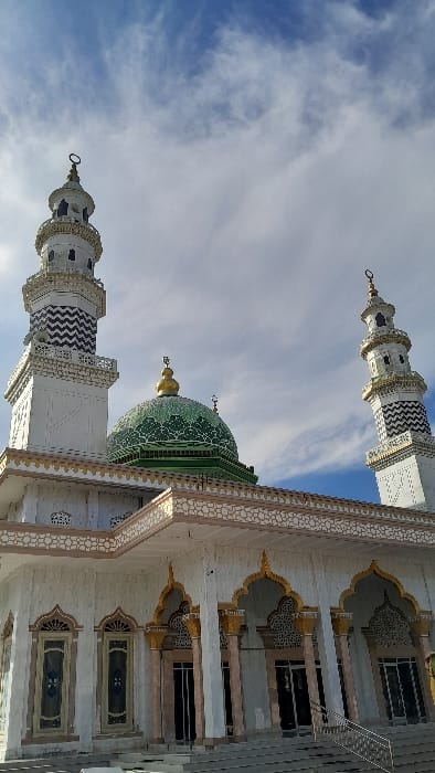 The image displays the exterior of a mosque in Alueiemirah, Indonesia. The structure features a large, central green dome with intricate light-colored patterns and a golden finial at its apex. Two tall, white minarets, adorned with black and white chevron-patterned bands and crowned with golden finials, flank the dome. The main building facade is white, embellished with decorative light brown and gold geometric patterns along the roofline and surrounding multiple arched windows or entrance portals. These portals contain dark-tinted glass. A set of stairs with a silver-toned metal handrail leads up to the building's entrance. The scene is illuminated by daylight, under a blue sky partially covered with white, wispy clouds. No individuals, actions, or interactions are visible.