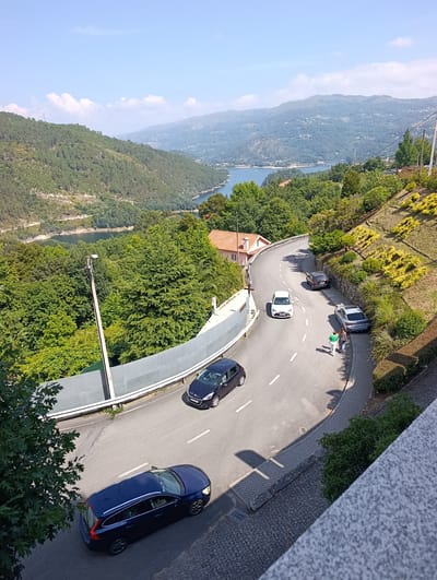 This aerial, wide-angle view captures a scenic, winding road in Terras de Bouro, Portugal, flanked by lush green trees and steep, terraced hillsides. The road curves around a hillside, with several cars parked and driving on it. In the foreground, a dark blue station wagon is parked on the lower part of the road. Further up, a dark colored SUV is parked, and a silver sedan is parked next to it. Two people, a woman in a green shirt and a woman in a black and white dress, are walking on the sidewalk next to the silver sedan. A white sedan is driving uphill. In the background, a wide river or lake is visible, with rolling, tree-covered hills in the distance. The sky is clear and blue with scattered white clouds. The environment appears to be rural and natural, with a focus on the road and its surroundings.