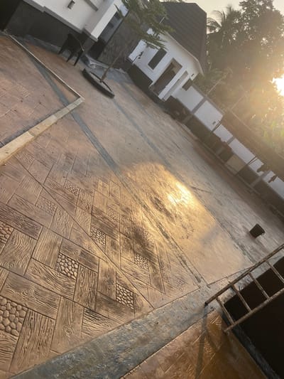 The scene depicts an outdoor paved courtyard or driveway in Ogbunu, Nigeria, under bright sunlight, likely morning or late afternoon, as indicated by the intense glare on the surface. The ground is covered with stamped concrete featuring reddish-brown and tan sections mimicking wood grain and various pebble patterns, interspersed with darker grey or blue linear dividers. In the background, two modern, multi-story structures are visible; they feature white upper walls, dark grey lower sections, and dark, sloped roofs. A small, sparsely leafed tree is planted in a black tire, positioned on the paved surface near the buildings. A black plastic chair rests near the base of one building. To the far right, a segment of a black metal railing is present in the foreground, and a small black bucket or cup lies on the paved surface further back. Dense green foliage, including palm trees, rises above a perimeter wall in the distant background. No individuals or active subjects are present.