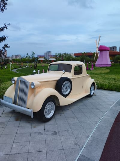 In Jiguan District, China, a cream-colored vintage coupe is parked on a tiled pathway in a park. The car features a prominent chrome grille, dual headlights, and a spare tire mounted on the passenger side. In the background, a vibrant pink windmill with illuminated blades stands out against a backdrop of greenery and city buildings. Several individuals are visible walking and standing in the park, which includes landscaped gardens and pathways. The sky is overcast with a pale blue hue.