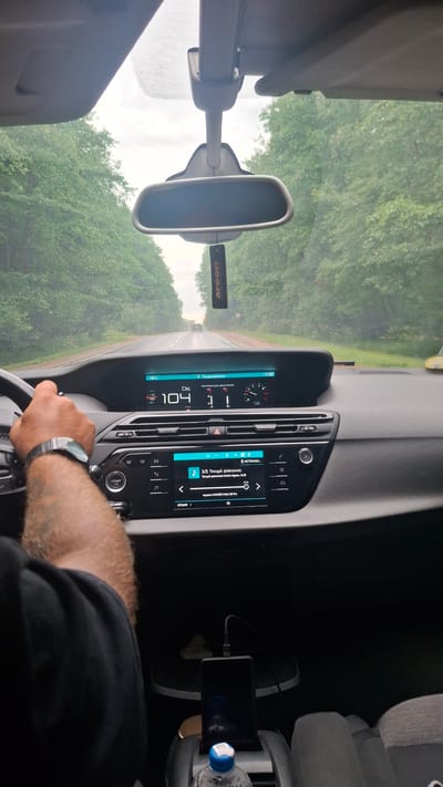 The image shows the interior of a car driving on a road lined with trees. The view is from the driver's perspective, with a hand on the steering wheel. The car's dashboard displays information on a digital screen, including speed and engine temperature. A phone is mounted in the center console and is charging. The weather appears overcast, with some moisture on the windshield. A car air freshener hangs from the rearview mirror. The location is identified as Khachenchitski Sel'skiy Sovet, Belarus, implying a rural or semi-rural setting.