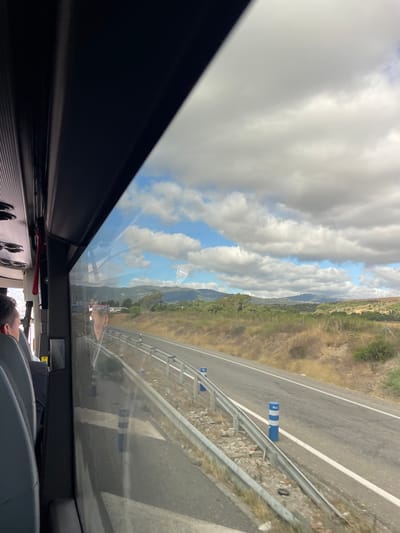 The image shows a view from inside a vehicle, likely a bus, looking out through a large window. The scene is a highway with a guardrail and blue and white traffic cones visible along the side of the road. Beyond the road, there is dry, brushy terrain and rolling hills with some greenery. In the distance, mountains can be seen under a partly cloudy sky. The clouds are large and white, with patches of blue sky visible between them. The sun appears to be shining, suggesting it is daytime. The reflection on the window shows a person's head and shoulder, implying they are a passenger. The overall impression is one of travel on a sunny day, with a scenic view of the Spanish countryside.