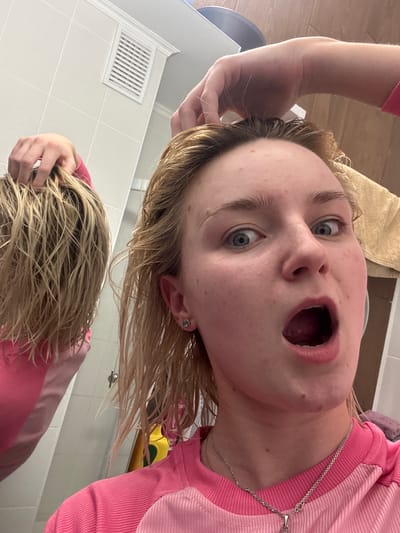 A young individual with light blonde, wet hair and wide blue eyes is the central subject of this close-up, rotated image. They are wearing a pink ribbed long-sleeved shirt and a silver chain necklace, with a small silver earring visible on their left ear. Their mouth is open wide, conveying an expression of surprise, awe, or playful shock.
The person appears to be looking into a mirror, evidenced by a distinct reflection of their wet hair and upper torso, including part of their pink shirt, in the lower section of the frame. One of their hands is visible touching their wet hair on the left side of their head, and a reflected hand is also seen in the lower part of the image, similarly touching hair.
The setting is an indoor space, likely a bathroom, indicated by white tiled walls with visible grout lines and a white rectangular air vent/grille mounted on the wall. A beige towel is draped in the upper right background. Partially visible objects in the reflection include yellow and green items, and a metal fixture that could be part of a sink or shower. The unusual, low-angle perspective, combined with the image's rotation, creates a dynamic and somewhat disorienting view, suggesting the photo was taken while looking upwards, possibly from a sink or bath. The wet hair implies recent washing. No visible text is present in the image. The location context is Ligolambi, Russia.