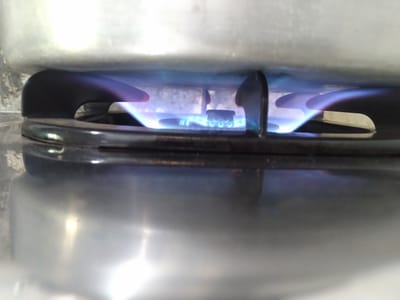 The image depicts a close-up, low-angle view of a lit gas stove burner with a blue flame, located in Gampongbaro, Indonesia. A large, silver-colored metal pot or cooking vessel is positioned directly above the burner, partially obscuring the top portion of the flame and the rear of the stove. The black metal grates of the stove support the pot, with two distinct support arms visible. The bright, steady blue flame emanates from the circular burner, indicating efficient combustion. The foreground shows the highly reflective, polished surface of the stove cooktop, which mirrors the blue flame and ambient light. The background is out of focus, suggesting a light-colored wall or backsplash behind the cooking area. No people are visible in the scene.