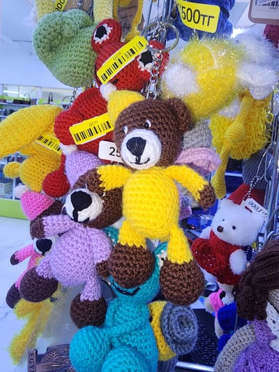 This image shows a close-up, eye-level view of a display of crocheted stuffed animals and keychains, likely in a retail environment. The items are arranged in a dense cluster, hanging from a rack or similar fixture. Prominent among the objects are several teddy bear keychains, all crocheted. One large yellow and brown teddy bear is centrally located, with its arms outstretched. Other smaller teddy bears in various colors, including white with a red scarf and hat, and purple, are also visible. Some of the items have yellow tags with barcode-like patterns and numbers, such as "0130" and "25".
The background is blurred, but suggests an indoor setting with shelving and other merchandise. There are no people visible in the frame, and no specific actions or interactions are depicted. The overall impression is one of a vibrant and colorful collection of handmade crafts.
The location is identified as Ushtobe, Kazakhstan.