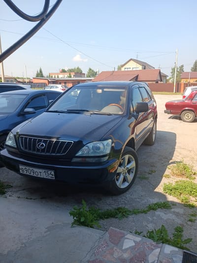 A black Lexus RX is parked on a dirt and concrete lot in Prokudskoye, Russia. The car has a license plate that reads "B909OT 154 RUS". A blue car is parked to the left of the Lexus. A red car is parked to the right of the Lexus. Several houses with red roofs are visible in the background, along with trees and a utility pole. The sky is blue and clear.
