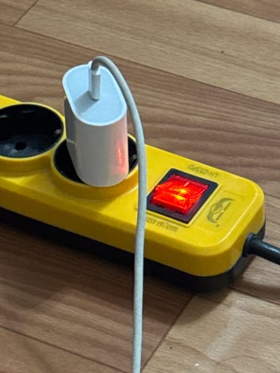 A white phone charger with a red indicator light is plugged into a yellow power strip. The power strip has a red illuminated switch and two black electrical outlets. The power strip is resting on a wooden floor. The image is a close-up shot, and there are no people visible. The scene is indoors.