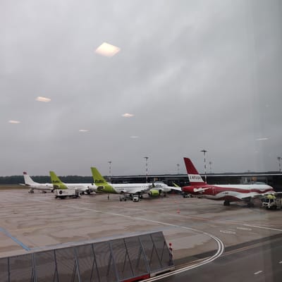 The image displays an airport apron scene viewed from an interior perspective through a window, under an overcast sky in Skulte, Latvia. The lower foreground is partially obstructed by a dark metal mesh fence. The tarmac, comprising both asphalt and concrete surfaces, is visibly wet with puddles, indicating recent rain.

Four passenger aircraft are parked in a line. The three leftmost aircraft share a white fuselage with vibrant green tailfins and engine nacelles, emblazoned with "Baltic" on the tail. The second aircraft from the left also features "airBaltic" in smaller script on its fuselage and the registration "YL-ABO". The fourth aircraft on the right exhibits a distinct red and white livery with "LATVIJA" prominently displayed on its red tailfin and "YL-CSL" visible on its fuselage near the nose. All four aircraft appear to be Airbus A220-300 models.

Multiple pieces of ground support equipment are active on the apron. A white "LSG Sky Chefs" catering truck is positioned adjacent to the leftmost aircraft. A yellow-green fuel truck is parked next to the "LATVIJA" aircraft on the far right. Numerous baggage carts and other utility vehicles are visible. Orange traffic cones delineate areas around the aircraft. Several ground crew personnel are performing tasks near the aircraft, notably around the second AirBaltic plane from the left.

The mid-ground reveals a dark-colored, modern airport terminal building featuring extensive glass panels. Tall light poles or antenna structures are scattered across the tarmac and near the terminal. The upper section of the image shows significant reflections of square and rectangular indoor lighting fixtures on the window surface, partially obscuring the uniformly grey, cloud-filled sky.