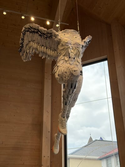 An artistic sculpture made of white Nike sneakers hangs from the ceiling in a modern building with wooden paneling. The sculpture depicts a winged humanoid figure in a dynamic pose, with its arms outstretched and legs extended. The figure is constructed entirely from disassembled sneakers, with the shoes forming the wings, torso, head, arms, and legs. Several Nike logos are visible on the sculpture. The sculpture is illuminated by spotlights from above and is positioned in front of a large window that looks out onto a cloudy sky and the rooftops of buildings in a city. Below the window, part of a tiled roof and a building with windows are visible. The overall impression is one of an avant-garde art installation. The specific location in Roermond, Netherlands, is the Nike Factory Store Roermond.