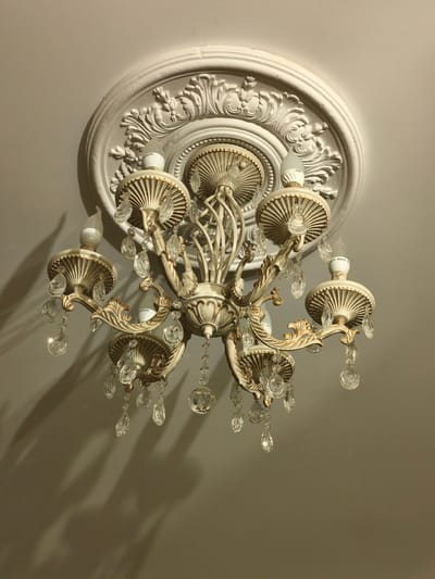 This is a close-up, low-angle, medium shot of a vintage-style chandelier hanging from a white, ornate ceiling medallion. The chandelier is made of a light-colored metal, possibly brass or a painted finish, with intricate scrollwork and ribbed shades. Several clear glass or crystal teardrop-shaped prisms are suspended from the arms of the chandelier, catching the light. The lightbulbs themselves appear to be modern LED bulbs, some of which are partially visible and labeled with Chinese characters. The ceiling is a plain, off-white surface. Shadows from the chandelier and its prisms are cast on the ceiling below, indicating a light source from above and to the right. There are no people or animals present in the image. The setting appears to be indoors, in a residential or possibly a public space with a decorative ceiling.