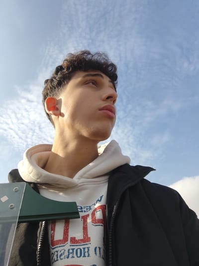 A low-angle outdoor portrait depicts a young male subject looking upwards and to the right against a clear blue sky with scattered white, rippled clouds. The subject, with light skin and dark curly hair featuring a faded cut, is illuminated by sunlight on his right ear and the right side of his face. He is wearing a light-colored hooded sweatshirt with a partially visible red and blue graphic print, layered under a dark-colored, partially unzipped jacket. In the lower-left foreground, a portion of a clear panel, secured by fasteners, is visible, partially obscuring a dark green angular object attached to it. The scene is located in Mankat al-Hatab, Syria.
