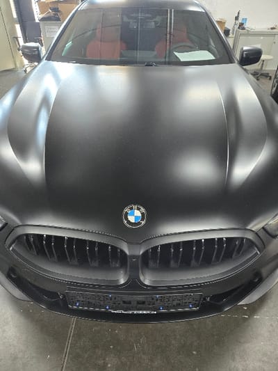 The image displays the front view of a matte black BMW 8 Series coupe, parked indoors, likely within a commercial garage or workshop. The vehicle features a large, black vertical-slat kidney grille and a prominent BMW emblem centered on the hood. A black license plate holder is affixed to the front bumper, displaying "KFZ MEISTERBETRIEB" and what appears to be "Sprendlingen" or "Dreieich-Sprendlingen" on a blue strip, indicating a workshop or dealership in Sprendlingen, Germany. Through the windshield, red leather interior seating and a white document on the dashboard are visible. The surrounding environment includes a dark floor, with the background revealing stacked cardboard boxes, an office chair, and a labeled white container on the left, and white cabinets, a workbench, and various tools or equipment on the right. Overhead artificial lighting is reflected on the car's surface. No individuals are present, and no actions or interactions are depicted.