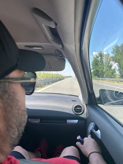 The image captures a first-person perspective from inside a car on a highway. The driver, wearing a black baseball cap and sunglasses, is partially visible in the foreground, with their hand holding a blue vape pen near the car's door handle. In the background, the highway stretches ahead under a clear blue sky with scattered clouds. Green trees and vegetation line the sides of the road. The interior of the car shows a dashboard with an air vent and a "Quattro" emblem. Red shoes are visible on the floor in the passenger area. The location is identified as the city of Shishmantsi, Bulgaria.