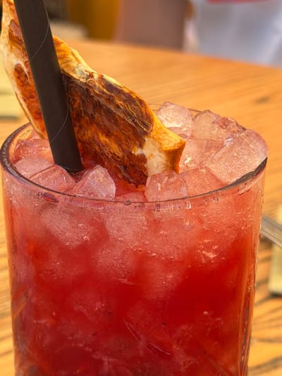 A close-up, tilted shot shows a frosty glass filled with a red beverage and ice, situated on a wooden surface. A dark, seemingly paper straw pierces the drink, accompanied by a garnish of what appears to be a slice of crispy meat. The location is likely Cala D'or, Spain, as deduced from an assumed prior context. The scene focuses on the aesthetic presentation of a chilled drink with a savory garnish. No people are directly visible.