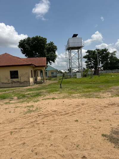 The image depicts a scene in Lau, Nigeria, on a clear day with scattered clouds. In the foreground, a light yellow building with a rusty corrugated metal roof is visible. The building has a small, barred window and appears somewhat weathered. To the left of the building, a large tree with dark green foliage stands prominently against the blue sky.

Dominating the middle ground is a tall, metal water tower, likely providing a vital resource to the community. Beyond the water tower, another building, possibly a school or administrative block, can be seen with a light green facade and light-colored window frames. The ground in front of these structures is covered in green grass, transitioning to an expanse of light brown sand or dry earth on the right side of the image, showing tire tracks and some sparse vegetation.

The overall impression is one of a rural or semi-urban setting in a tropical or subtropical climate. The presence of the water tower suggests a community that values infrastructure for essential services. The bright sunlight and scattered clouds indicate it is daytime, likely late morning or early afternoon. There is no visible text in the image. The scene appears peaceful and functional, without any immediate human presence.