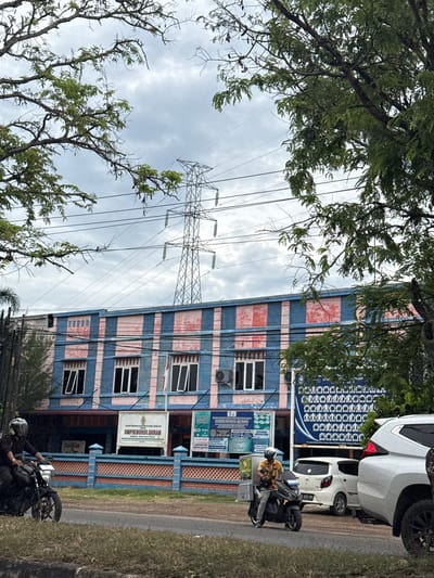 In Meunasahmanyet, Indonesia, under a cloudy sky, a low-angle shot captures a street scene. A large, blue and pink striped building with multiple windows and air conditioning units dominates the mid-ground. Above the building, a tall electricity pylon with power lines stretches across the sky. In the foreground, two motorcycles are visible on the paved road. The first motorcycle, on the left, is ridden by a person wearing a black shirt and a white helmet. The second motorcycle, on the right, is a scooter with a cargo box attached to the rear, ridden by a person in a yellow shirt and white helmet. A white car is parked behind the second motorcycle, and partially visible on the far right is the front of a white SUV. Trees with green foliage frame the top and sides of the image, partially obscuring the sky and the building. Signs on the building include "SMPI NURULQURAN".