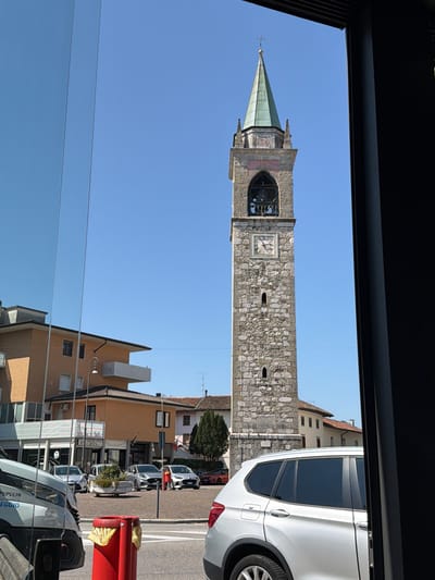 The image shows a street scene in the city of Udine, Italy, on a clear, sunny day. In the background, a prominent stone bell tower with a green steeple dominates the skyline. The tower has a clock face, arched openings, and is constructed of rough-hewn stones. To the left of the tower, several buildings are visible, including a light brown, multi-story building with balconies and windows, and a lower, darker building that appears to be a shop with signage. In the foreground and mid-ground, cars are parked and driving on a paved road. A silver SUV is parked on the right side of the image. A white van or truck is on the left, with some text visible on its side reading "PUPULIN GROUP LEGGI." A bright red trash bin with yellow bags is positioned on the road. The overall impression is of a typical urban street with historical architecture.