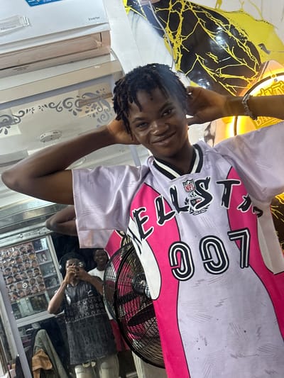 A young man with braided hair stands indoors, wearing a lavender jersey with pink accents, the words "EST. LOGO" visible on the front, and an NFL logo patch on the chest. His right hand is raised near his face. Behind him is an electric fan, and reflected in the glass behind him is another person wearing a black shirt with patterns. The background suggests a shop setting with various items displayed behind the reflected person. The location is Ogwashi-Uku, Nigeria.