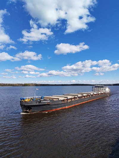 The image depicts a long, dark barge labeled "КУНЦЕВО" (Kuntsevo) navigating a body of water, likely a river or large lake. The barge is loaded with rectangular cargo, possibly concrete blocks. The sky above is bright blue with scattered white cumulus clouds. In the distance, a thin line of trees marks the horizon. The water surface is rippled, suggesting a light breeze. While the image suggests a waterway in Russia, it doesn't provide specific evidence it's in Pruzhinino. More information is required to place this scene in Pruzhinino, Russia.