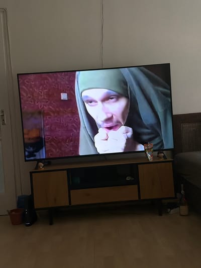 An indoor scene shows a large television screen displaying a close-up of a person's face, wearing a green head covering. The person's mouth is slightly open, and one hand is held near their chin. The television is placed on a wooden entertainment stand with drawers. To the left of the stand, there is a red bucket and a black electronic device with lights. To the right of the stand, on the television screen, a juice box is visible. A small plush toy and a bottle are also on the right side of the entertainment stand. The background features a red patterned wallpaper and a portion of a dark-colored couch. The scene is dimly lit, and the camera is positioned at a slight downward angle. There is no information available to suggest this image is located in Kalocsa, Hungary.