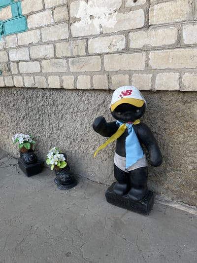 A black-painted statue, resembling a stylized child or anthropomorphic figure, stands on a cracked concrete pavement against a building wall in Synelnykove, Ukraine. The figure wears a white baseball cap with a yellow brim and a red New Balance (NB) logo, a yellow ribbon tied around its neck, a light blue fabric piece resembling a tie, and striped fabric around its waist. Its right arm is raised in a waving gesture. To the left of this central figure, two smaller black statues, appearing to be stylized crawling figures, are positioned in a line. Each small statue holds a small pot containing artificial white flowers with green foliage. The wall behind the figures consists of a rough grey stucco base, topped by a light-colored brick section with visible weathering, peeling white paint/plaster, and some turquoise paint remnants on the far left. Faint red markings and other graffiti are visible on the brickwork.