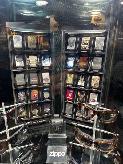 The image shows a display case inside a store, likely in Lake Buena Vista, Florida, United States. The case contains numerous Zippo lighters, many of which feature Harley-Davidson logos and designs. A selection of eyeglasses on a wire display rack is also visible, partially obscuring the Zippo lighters. The overall environment appears to be a retail setting.