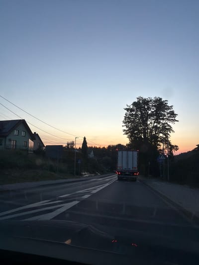 The image depicts a road scene at dusk in Bochnia, Poland. A large truck with its brake lights illuminated is traveling on a two-lane road. The road has white lane markings. To the left of the road, there are houses situated on a slope, and power lines are visible overhead. To the right of the road, a large tree stands prominently, and further down the road, there are more trees and some smaller structures. The sky is a gradient of pale blue at the top, transitioning to orange and pink hues near the horizon, indicating the setting sun. The overall atmosphere is tranquil and suggests the end of the day.