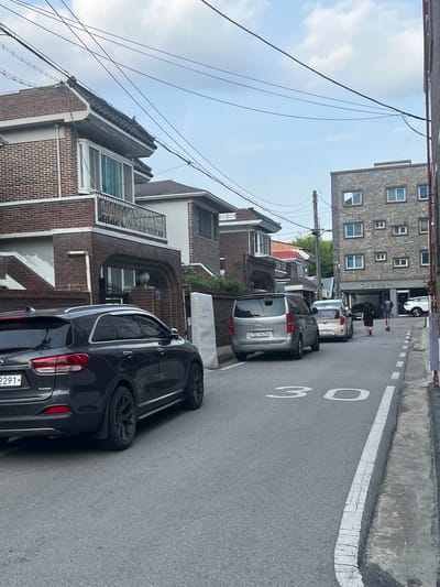 The image depicts a narrow urban street in Gwangju, South Korea, characterized by a mix of residential and commercial buildings. In the foreground, a dark gray Kia Sorento SUV is parked on the right side of the street, partially obscuring a license plate that ends in "2291". To the left, a silver minivan with license plate "765 9673" is also parked. Further down the street, a white sedan with license plate "205 7892" is visible.

A speed limit sign of "30" is painted on the asphalt road. Two individuals are walking in the distance on the right side of the street, with one on a scooter. The buildings present a variety of architectural styles, including a brick house with a traditional tiled roof on the left, and a modern gray brick building with numerous square windows on the right. Power lines crisscross overhead. The sky is partly cloudy.