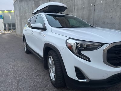 A white GMC Terrain SUV, equipped with a roof cargo box, is parked on an asphalt road in front of a concrete wall. The vehicle is positioned at an angle, with its front right side closer to the camera. The SUV features a dark-colored tire with a silver rim on the visible front wheel. To the left of the vehicle, a building with a light-colored facade and green accents is visible, along with yellow bollards. The background consists of a rough, grey concrete wall. The lighting suggests it is daytime, possibly overcast. The scene appears to be located in Valdemoro, Spain, based on the context provided.