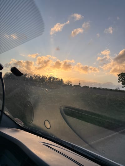 The image shows a view from inside a vehicle, likely a car, looking out towards a sunset. The sky is visible with light, fluffy clouds illuminated by the setting sun, casting a golden glow. Sunbeams are visible through the clouds. A tree line is on the horizon. The vehicle's dashboard and windshield are in the foreground. The location appears to be Ruyton-XI-Towns, United Kingdom, though there's no specific evidence in the image to confirm that.