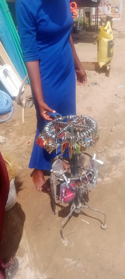 The image captures an outdoor commercial scene in Gauraka, Nigeria. A person, partially visible, wears a solid blue dress and stands barefoot with their right foot on dry, light-brown soil. Their left hand grips a blue padlock hanging from a metal display stand.
The display stand is a two-tiered, dark-colored metal structure with a tripod base. The upper tier presents numerous padlocks of various colors (red, blue, green, silver) and sizes, each suspended by its shackle with an accompanying key. The lower tier displays blister-packaged items, notably a red "X-FORCE" branded product.
In the left background, a stack of teal-colored corrugated plastic sheets leans vertically. Below these, a white ceramic squat toilet pan, coiled blue hoses, and a light-colored wooden tool lie on the ground. The right background features a large yellow sack, branded "FEEDS" with a chicken logo, resting on a small wooden stand. Behind it, a dark bucket and potted plants are visible. Further back, a commercial sign partially reads "NOW SELLING" and displays prices, with large orange scissors hanging nearby. A second foot, clad in grey sandals, is partially visible in the extreme lower-left foreground.