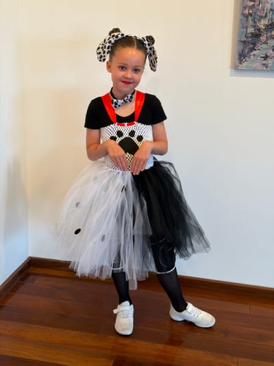 This image, captured in Ventspils, Latvia, depicts a young Caucasian female child, approximately 7-10 years old, standing centrally in an indoor setting. She is dressed in a costume featuring a black short-sleeved shirt, a white mesh top with a prominent black paw print on the chest, and red shoulder straps. A black and white polka-dot bow tie is worn with a black choker. Her headwear consists of a headband with black and white spotted dog ears. Her lower attire comprises a split tutu skirt, half white with scattered black circular spots and half solid black, worn over black tights or leggings. She wears white athletic shoes with velcro closures. Her hair is styled in two buns, and she has red lipstick applied, looking directly at the camera with a slight smile. The background features plain, light-colored walls and a polished dark reddish-brown wooden floor with visible grain, laid horizontally. A wooden baseboard runs along the bottom of the walls. In the upper right background, a framed abstract painting with cool tones is partially visible on the wall.